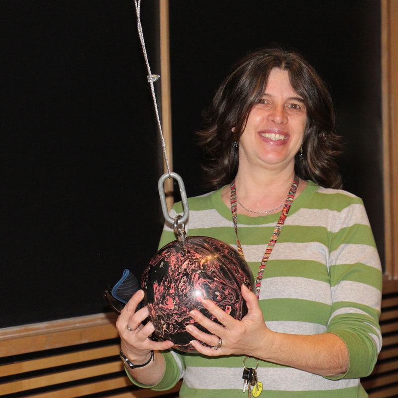Woman standing, wearing a striped shirt from the waist up holding a bowling ball suspended on a cable. 